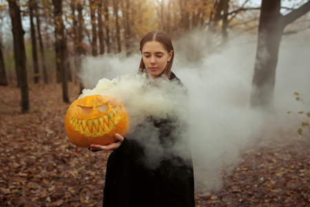 A beautiful girl with long hair in a black cloak is holding an orange pumpkin from which smoke is coming. Happy Halloween. dark autumn forest. horrors.の写真素材