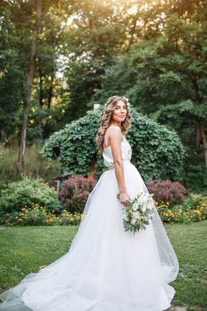 A cute curly woman in a white wedding dress with a wedding bouquet and wreath in her hair standing back to the camera in nature. Concept escaped bride. Forward to a happy bright future Runaway.の写真素材