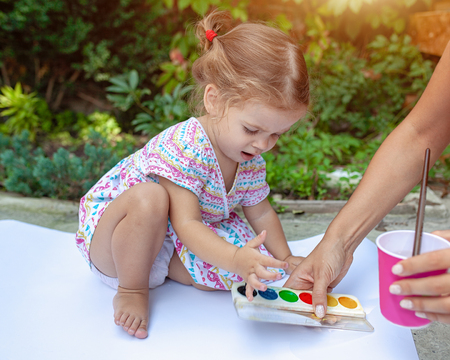 A little girl with curly hair sits on the floor and paints brightly colored paints on a large white sheet of paper. Summer vacation. Leisure for kids.の写真素材