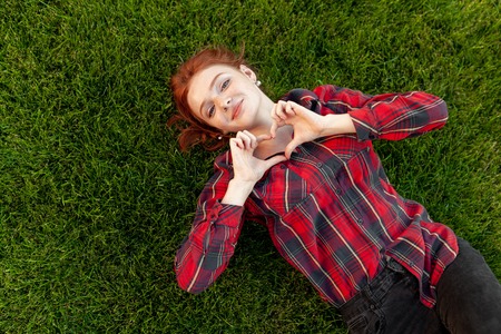 Beautiful red-haired young student with freckles lying on her back on the grass top view. Dressed in a red checked shirt, reveals a heart of fingers Love and positive.の写真素材