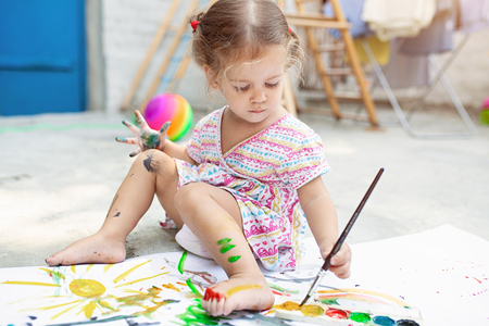 A little girl with curly hair sits on the floor and paints brightly colored paints on a large white sheet of paper. Summer vacation. Leisure for kids.の写真素材