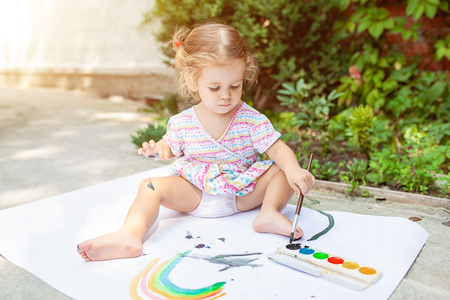 A little girl with curly hair sits on the floor and paints brightly colored paints on a large white sheet of paper. Summer vacation. Leisure for kids.の写真素材