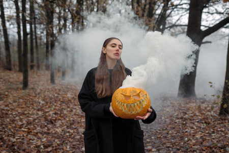 Young woman in the black coat holding the halloween pumpkin with the white smoke coming from inside of it in the autumn forestの写真素材