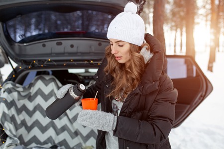 Young woman in woolen hat and black jacket stand neartrunk of the car and holds a cup of hot tea in her hands.の写真素材