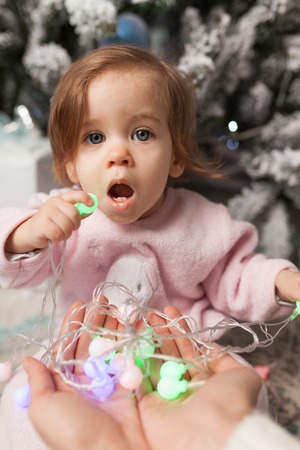 Small girl in the pink pajama sitting near the christmas tree playing with the garland.の写真素材