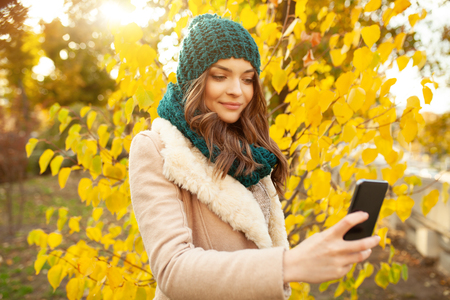 A beautiful young girl walks through the autumn park on the background of brightly colored leaves. Looks at your smartphone. Prints a message on the phone Social media everywearの写真素材