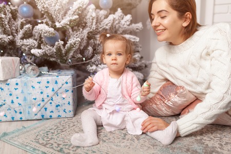 Happy New year Beautiful cute little girl sitting on the floor near the New Year tree next to her mother. Holiday mood, family values, childlike spontaneity and sweet atmosphereの写真素材