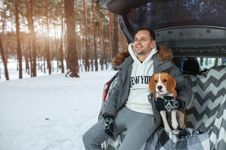 A young family of travelers a guy are sitting in the trunk of their car in an embrace with their friend dog Beagle. Winter forest on the background. Traveling with a dog by car.の写真素材