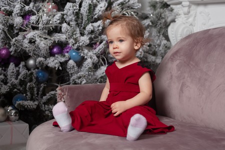 Merry Christmas and Happy New Year Beautiful cute little girl sitting on the sofa near the New Year tree. Holiday mood, childlike spontaneity and sweet atmosphereの写真素材