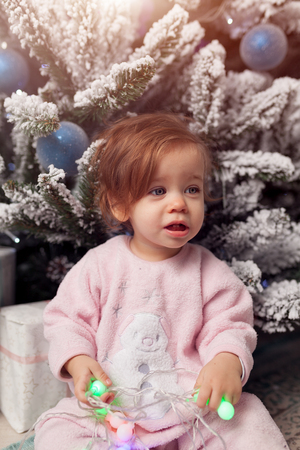 Small girl in the pink pajama sitting near the christmas tree playing with the garland.の写真素材