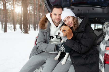 A young family of travelers a guy and a girl are sitting in the trunk of their car in an embrace with their friend dog Beagle. Winter forest on the background. Traveling with a dog by car.の写真素材
