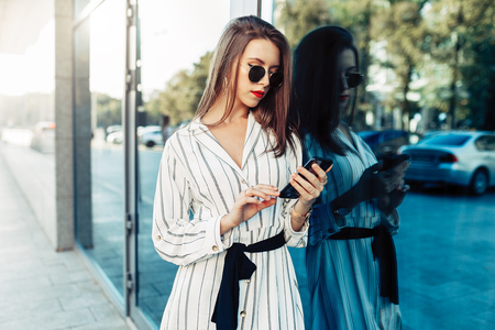Happy attractive young woman in sunglasses looking at smartphone screen while walking in the city. Dressed in stylish clothesの写真素材