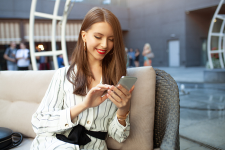 A young beautiful girl dressed in a striped fashionable dress is sitting in a summer outdoor cafe and is waiting for her order and her friends or her boyfriend. Prints a message on the phone.の写真素材