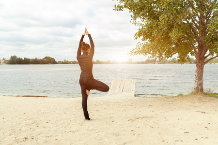 fitness, sport, people and lifestyle concept - young woman making yoga exercises on beach from back. rear view of a middle aged woman doing yoga outdoorsの写真素材