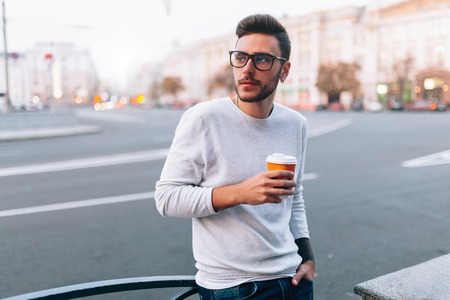 Hipster man standing with takeaway coffee, smiling plesantly, walking on th city street. Happy carefree handsome guy in eyewear. Millennial generationの写真素材