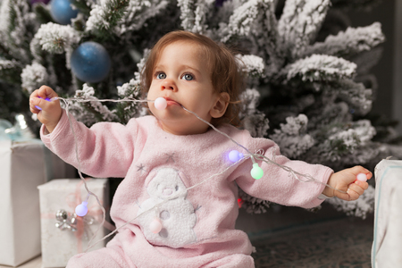 Small girl in the pink pajama sitting near the christmas tree playing with the garland.の写真素材