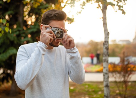 Young handsome hipster guy walks in a beautiful autumn park on the background of yellow leaves in warm sunny weather and takes pictures on a pig film camera. Autumn leisure time. Creative youth.の写真素材
