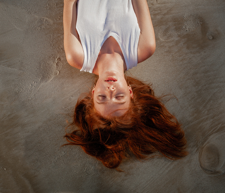 Young pretty red-haired woman in the white shirt laying on sand at the beachの写真素材