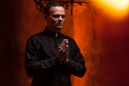 Young Catholic Praying priest. Portrait of priest Next to the candles prays with his hands folded near the face. Red wall on the background. Indoor handsome portrait of religion man.の写真素材