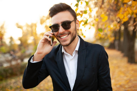 Handsome young stylish male businessman happy and smiling talking on the phone on a walk in the autumn park on the background of yellow leaves Dressed in a stylish business suit and sunglassesの写真素材