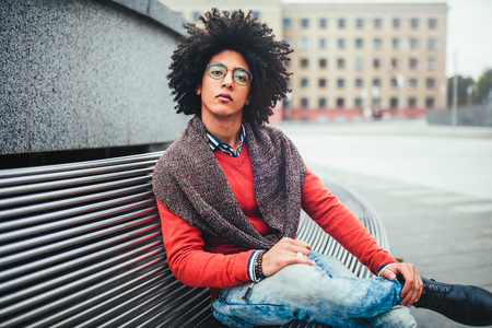 A handsome young Egyptian curly guy sitting on a bench dressed in a bright orange sweater and jeans. The student walks in the break between the lessons. Cute hipster guy.の写真素材