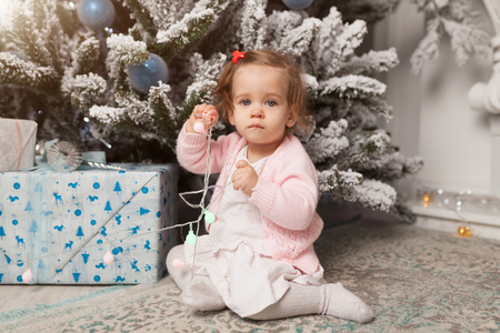 A little girl is playing with a garland with a New Year tree. Danger. Disobedient child. Good New Year spirit.の写真素材