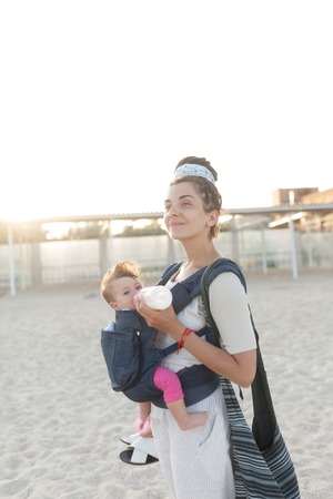 A small child sits in a backpack and walks along with the mother along the seashore. Summer family vacation concept. Woman with dreadlocks. Hipster. Modern motherの写真素材
