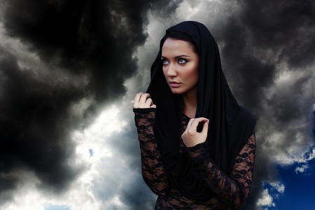 Young mysterious beautiful woman with the black hair and in the black hood and black transparent blouse at the sky backgroundの写真素材