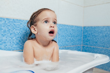 Fun cheerful happy toddler baby taking a bath playing with foam bubbles. Little child in a bathtub. Smiling kid in bathroom on blue background. Hygiene and health careの写真素材