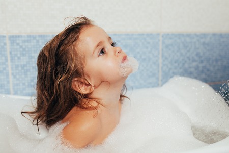 Fun cheerful happy toddler baby taking a bath playing with foam bubbles. Little child in a bathtub. Smiling kid in bathroom on blue background. Hygiene and health careの写真素材