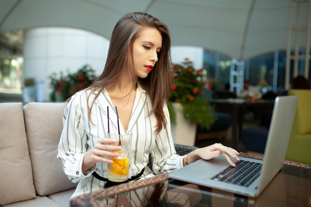 Businesswoman drink juice using wireless Internet connection on laptop and gadgets, messaging via popular social networks, dressed casually. People and technology concept.の写真素材