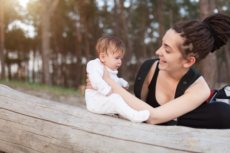 A beautiful smiling young brunette woman with long dreadlock hair hold a pretty baby. Mother looks tenderly at the child. They are in the green summer forest. With sunshine on the backgroundの写真素材