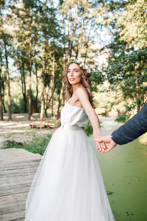 Follow me my love concept. Attractive young woman dressed in white wedding dress holding hand of her boyfriend and walking on the beautiful garden to happy futureの写真素材