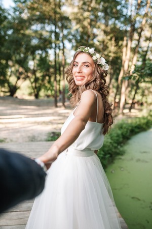 Follow me my love concept. Attractive young woman dressed in white wedding dress holding hand of her boyfriend and walking on the beautiful garden to happy future.の写真素材