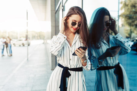 Happy attractive young woman in sunglasses looking at smartphone screen while walking in the city. Dressed in stylish clothesの写真素材