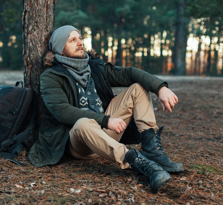 Pensive hipster guy wanderlust with touristic backpack enjoy tranquility during exploring nature environment, male hiker thinking about route to discover sitting on the groundの写真素材