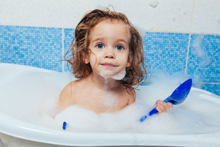 Fun cheerful happy toddler baby taking a bath playing with foam bubbles. Little child in a bathtub. Smiling kid in bathroom on blue background. Hygiene and health careの写真素材