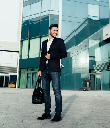 handsome young businessman with a beard and in a business suit standing on the street against the background of the office building next to a comfortable stylish leather bag. Business tripの写真素材