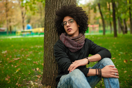 sad afro american guy with glasses sits in the park on the grass and looks into the camera. Stylish student with afro haircut.の写真素材