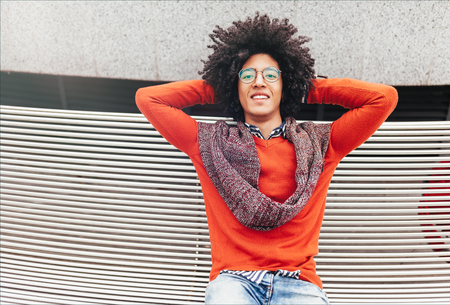 A handsome young Egyptian curly guy sitting on a bench dressed in a bright orange sweater and jeans. Happy people. A foreign student cupped his hands behind his head.の写真素材