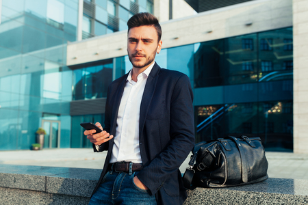 Young urban professional man using smart phone. Businessman holding mobile smartphone using app texting sms message wearing jacket With office buildings on the backgroundの写真素材