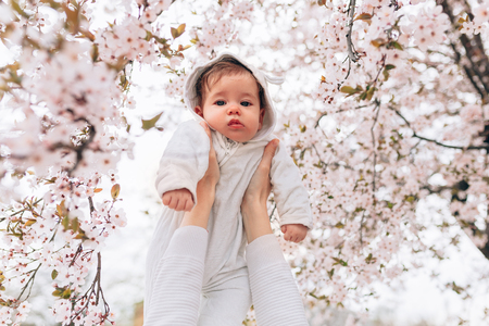 Portrait of happy joyful child in white clothes over tree flowers blossom background. Family playing together outside. Mom cheerfully hold little daughter Newborn spring conceptの写真素材