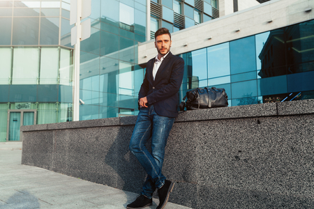 handsome young businessman with a beard and in a business suit standing on the street against the background of the office building next to a comfortable stylish leather bag. Business tripの写真素材