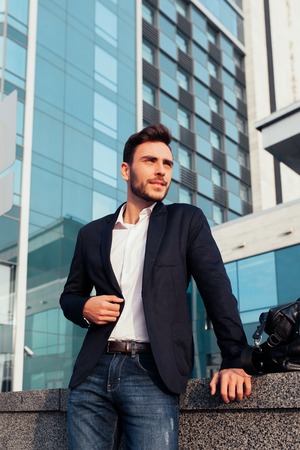 handsome young businessman with a beard and in a business suit standing on the street against the background of the office building next to a comfortable stylish leather bag. Business tripの写真素材