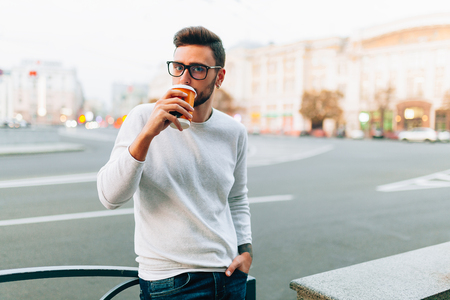 Hipster man standing with takeaway coffee, smiling plesantly, walking on th city street. Happy carefree handsome guy in eyewear. Millennial generationの写真素材
