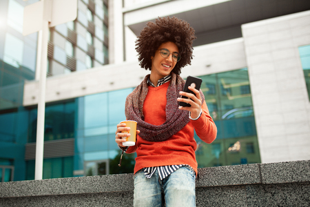 A handsome curly-haired guy of mixed race is dressed in a trendy youth-style scarf and sweater writes a message in a teleone against the background of office buildings. Youth culture.の写真素材