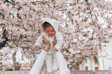 Portrait of happy joyful child in white clothes over tree flowers blossom background. Family playing together outside. Mom cheerfully hold little daughter Newborn spring conceptの写真素材
