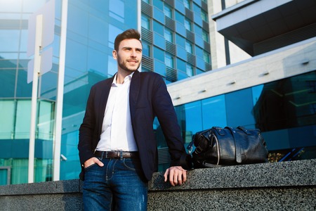 handsome young businessman with a beard and in a business suit standing on the street against the background of the office building next to a comfortable stylish leather bag. Business tripの写真素材