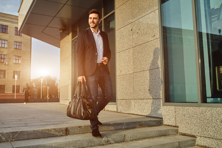 Men's fashion. A young male businessman in a trendy suit and a white shirt with a big black handbag comes out from a fashionable clothing store. Outdoor.の写真素材