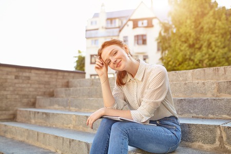 young beautiful student writes an essay in her notebook sitting on the steps stairs outdoor Red-haired girl with freckles study at free timeの写真素材